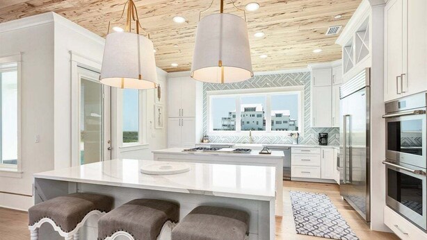 Una cucina moderna con mobili bianchi, una grande isola con sgabelli da bar, elettrodomestici in acciaio inossidabile e vista sugli edifici attraverso le finestre.