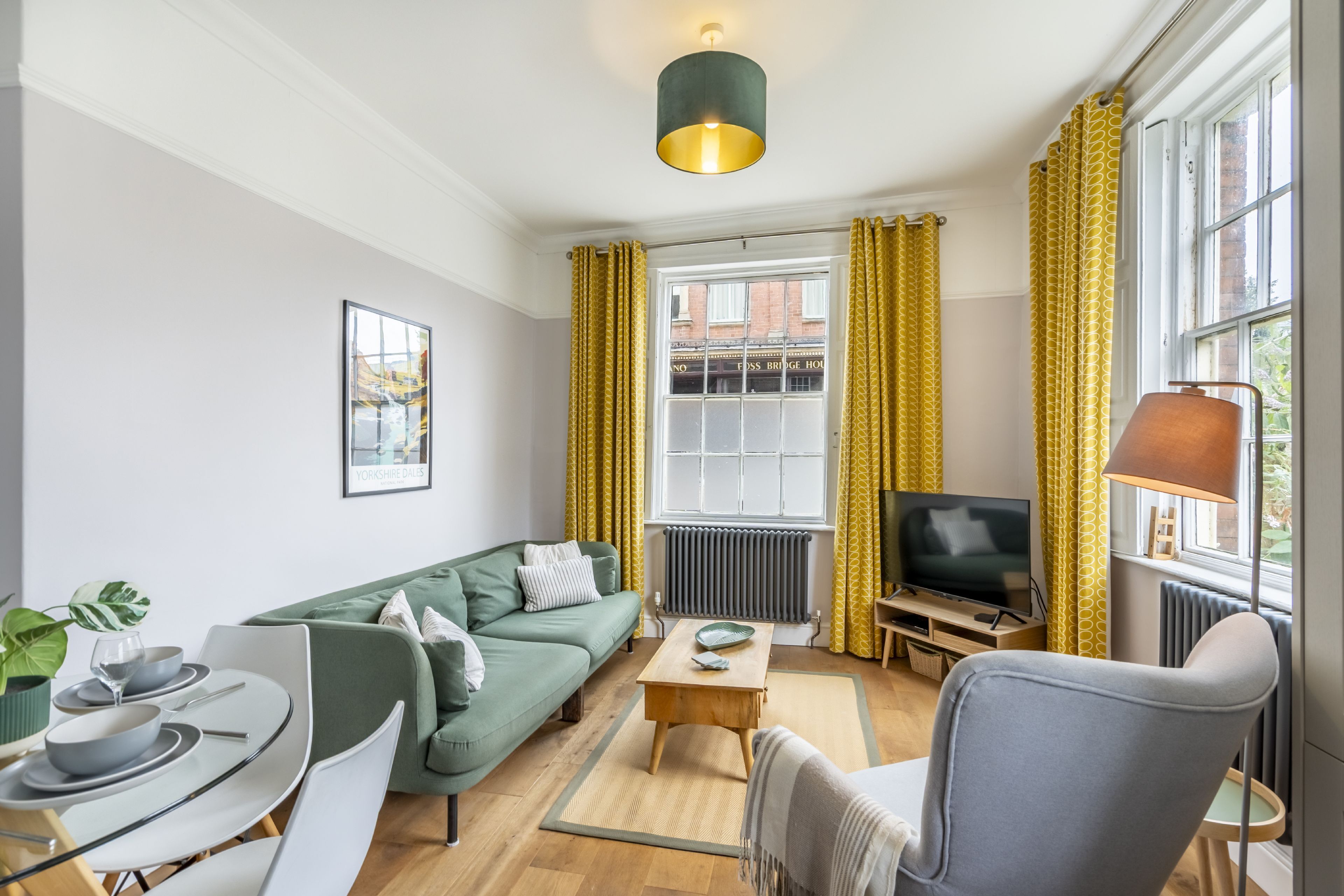 A modern living room with a green sofa, a dining table with chairs, a television, and yellow curtains.