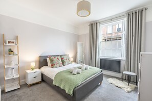 A bedroom with a bed, bedside tables, a radiator, a window with curtains, and a view of the street.