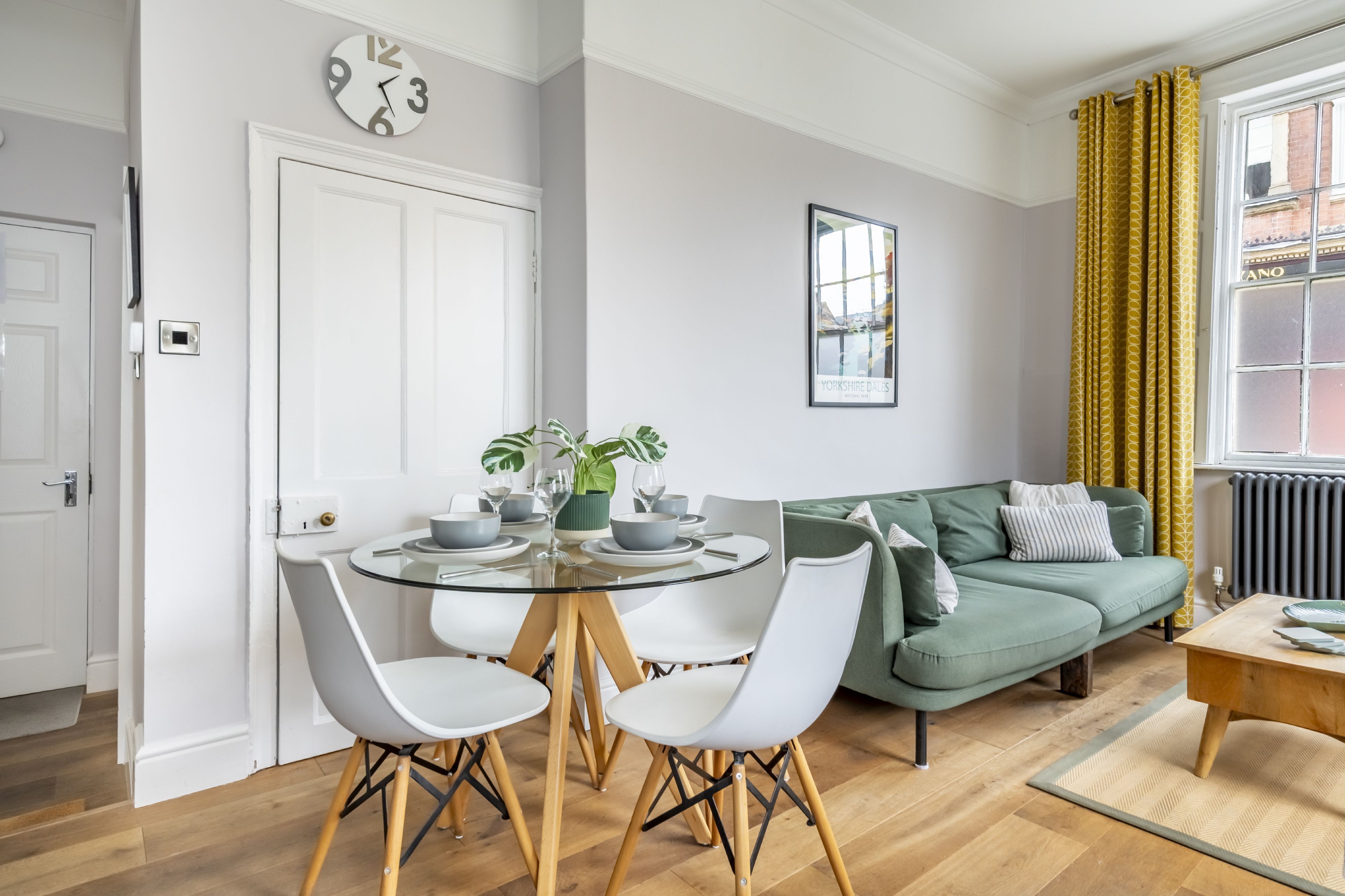 A modern living room with a round glass table, white chairs, a green sofa, and a framed picture on the wall.