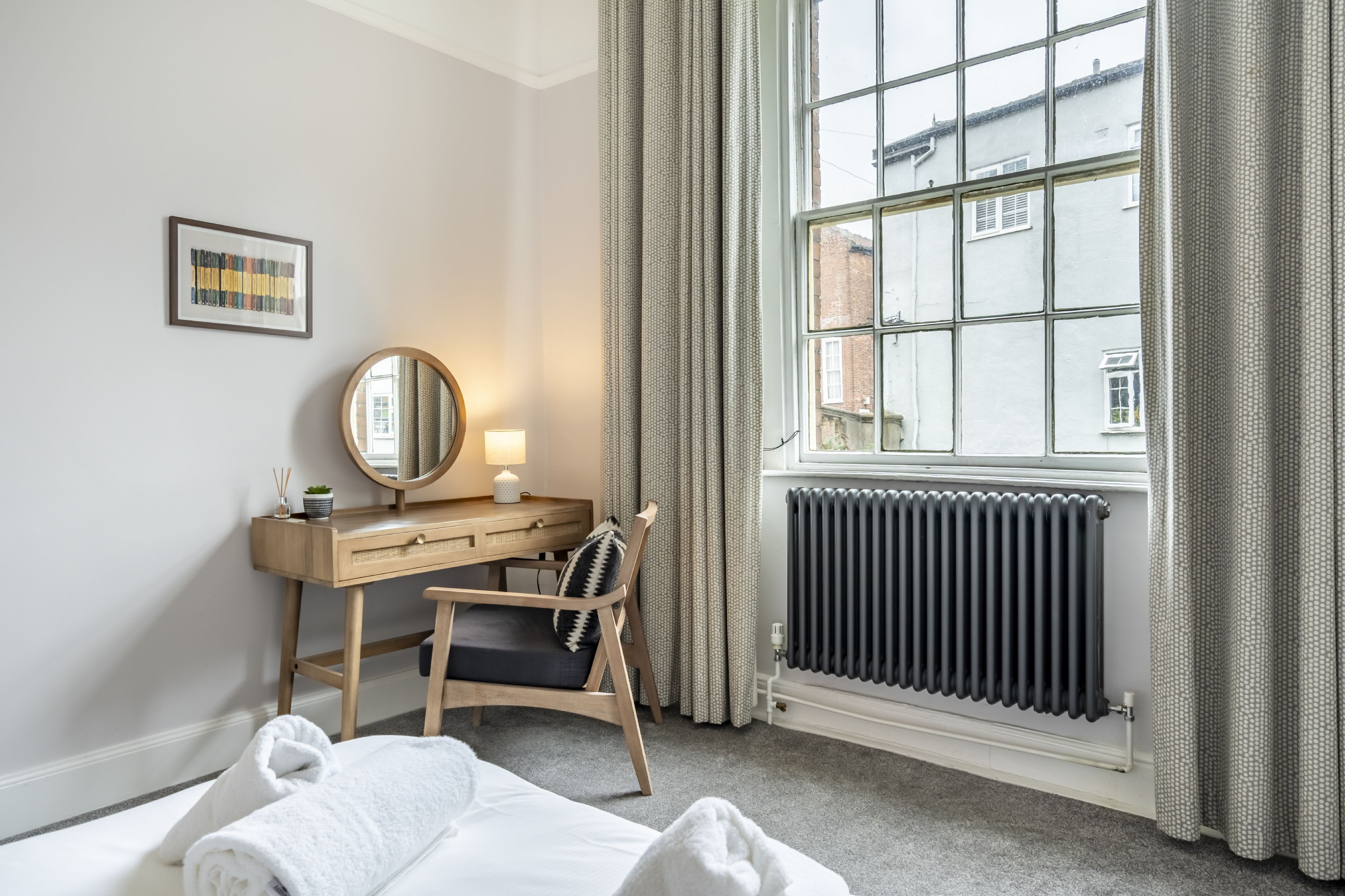 A hotel room with a bed, a desk with a chair, a mirror, a radiator, and a window with curtains.