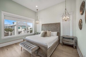 Een slaapkamer met een groot bed, een bankje, een nachtkastje en een raam met uitzicht op huizen.