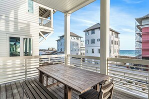 Een houten terras met een tafel en stoelen, uitkijkend op een strand met gebouwen op de achtergrond.