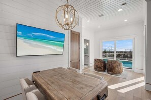 Een moderne eetruimte met een houten tafel, witte stoelen met kussens, een grote flatscreen televisie aan de muur en uitzicht op een zwembad door grote ramen.