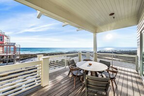 En overdækket terrasse med et rundt bord og stole, med udsigt over en strand og ocean.