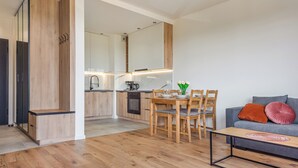 Un apartamento moderno con piso de madera, un área de comedor con mesa y sillas, una cocina con horno empotrado y pileta, y un sofá gris con cojines.