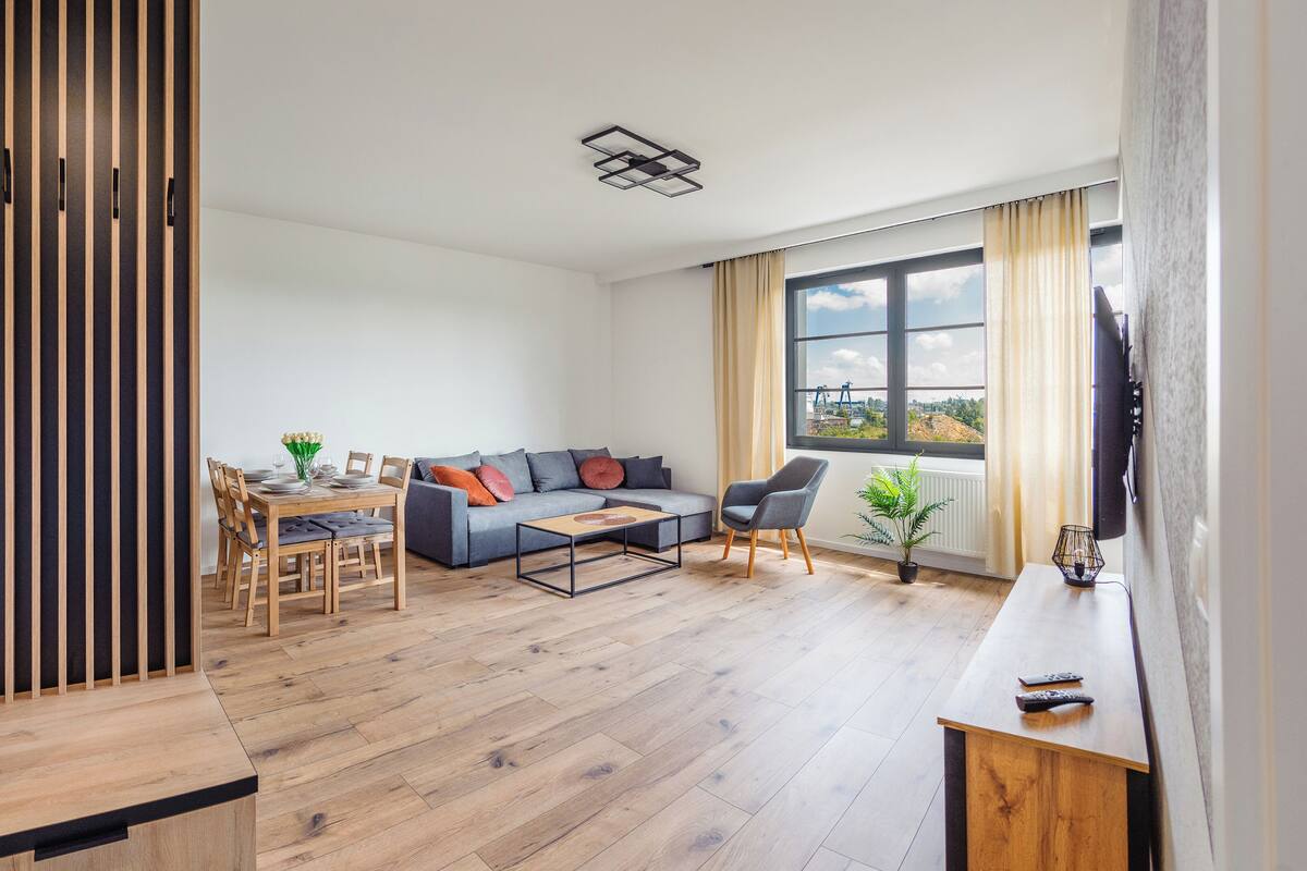 A modern living room with a dining area, a grey sofa, wooden flooring, and a large window with a view.