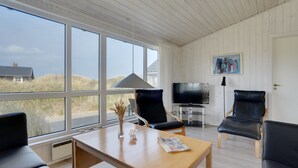 Ein Zimmer mit Holztisch, zwei schwarzen Ledersesseln, einem Fernseher und großen Fenstern mit Blick auf Dünen.