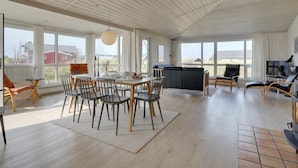 Ein geräumiges Zimmer mit Holzbalkendecke, großen Fenstern, einer Essecke mit Stühlen und Tisch sowie einer gemütlichen Sitzecke mit Sofa und Sesseln.