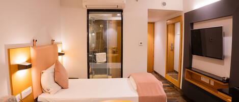 Habitación de hotel con cama, sofá, televisor, una mesita y baño con puerta de cristal.
