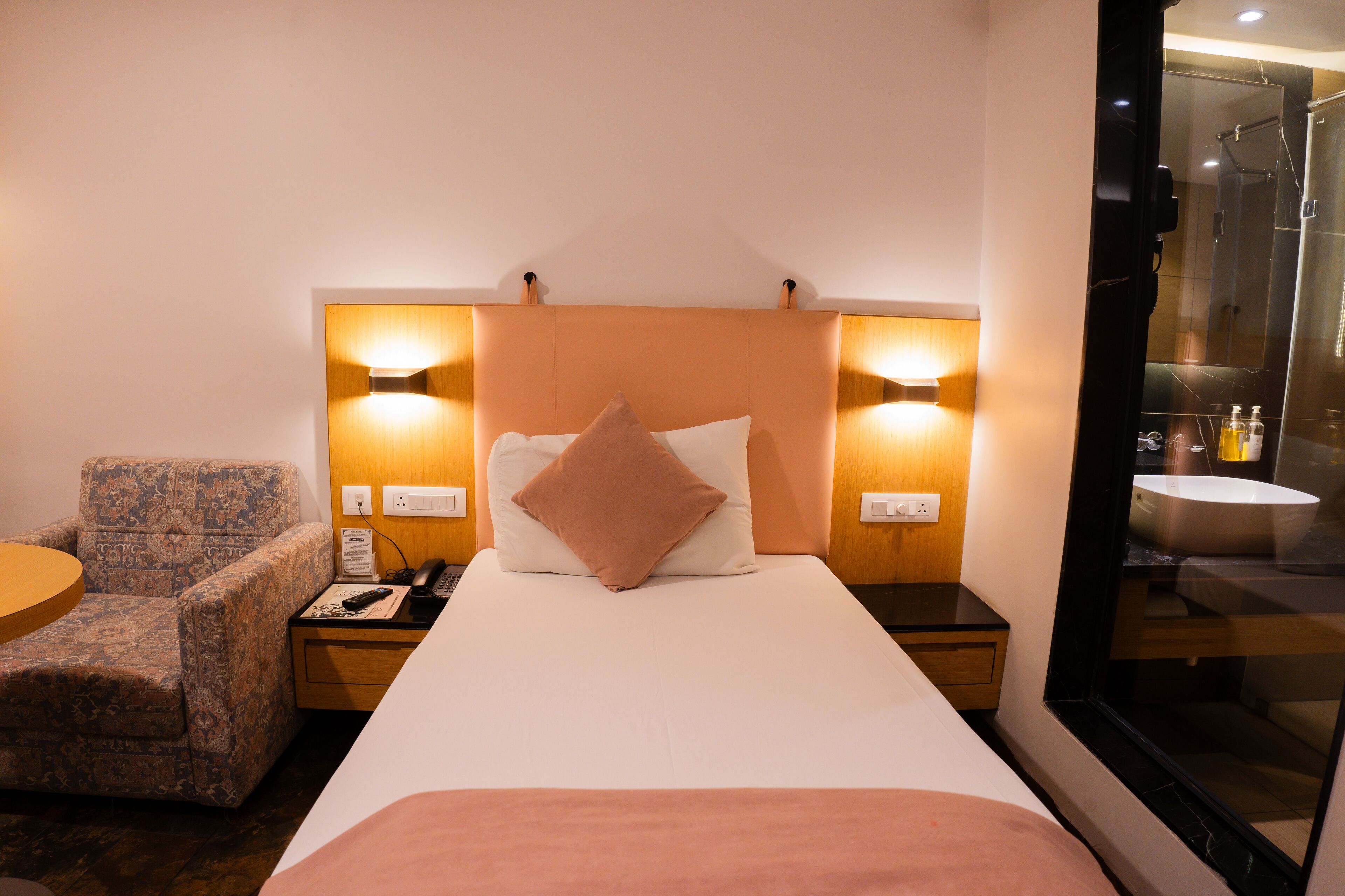 Une chambre d’hôtel avec un lit, une chaise, une table de chevet et une salle de bain visible à travers une porte en verre.
