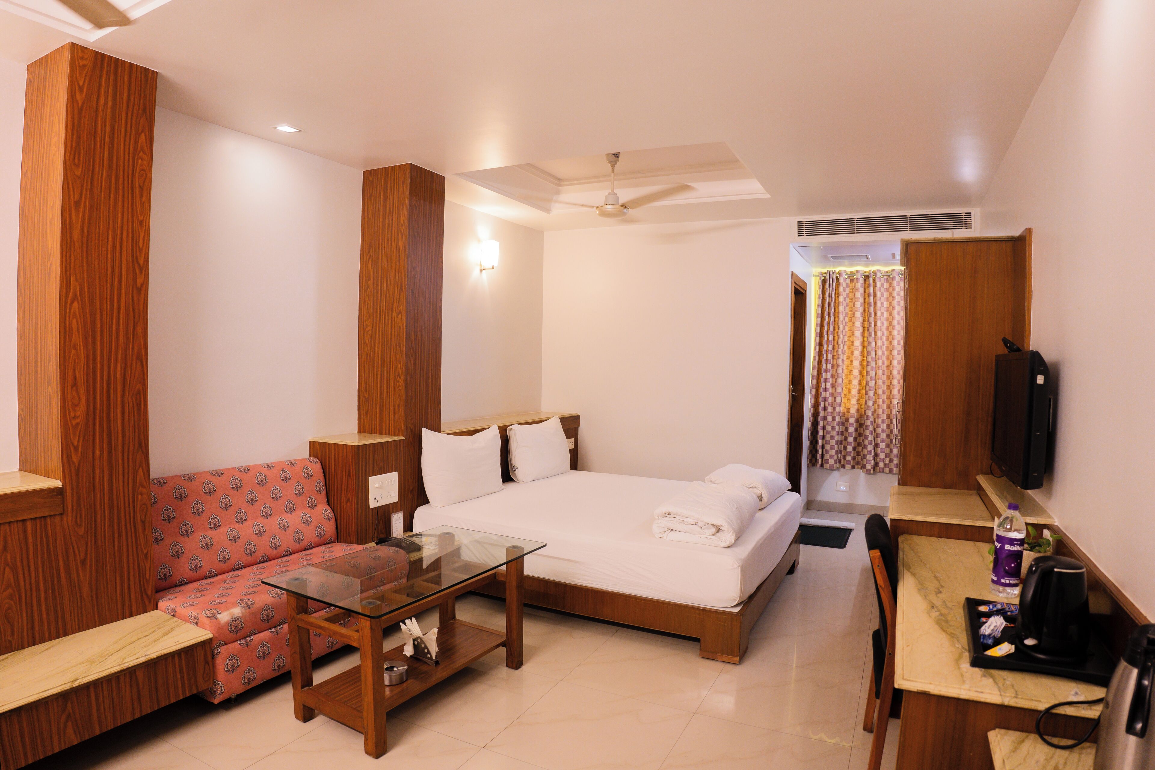A hotel room with a bed, a sofa, a glass-top table, a desk with a chair, and a television.