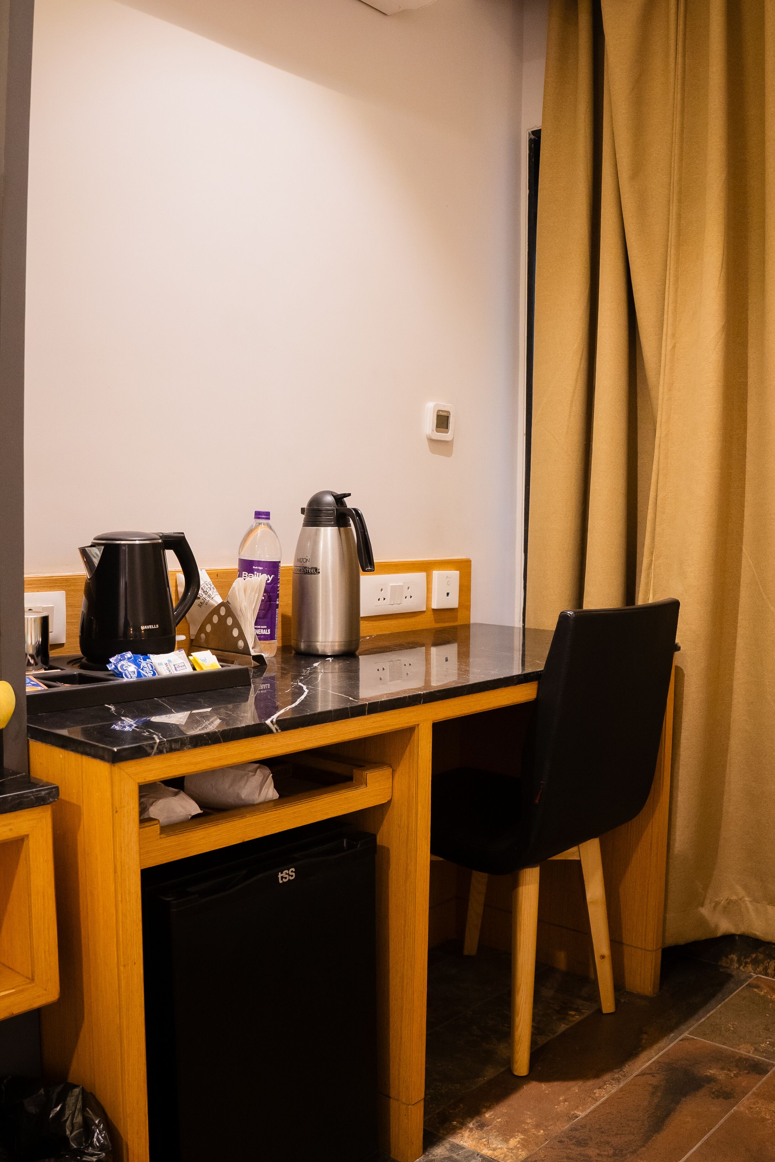 A compact hotel room with a wooden desk, a black chair, a compact refrigerator, a coffee maker, a bottle of water, and a power outlet.