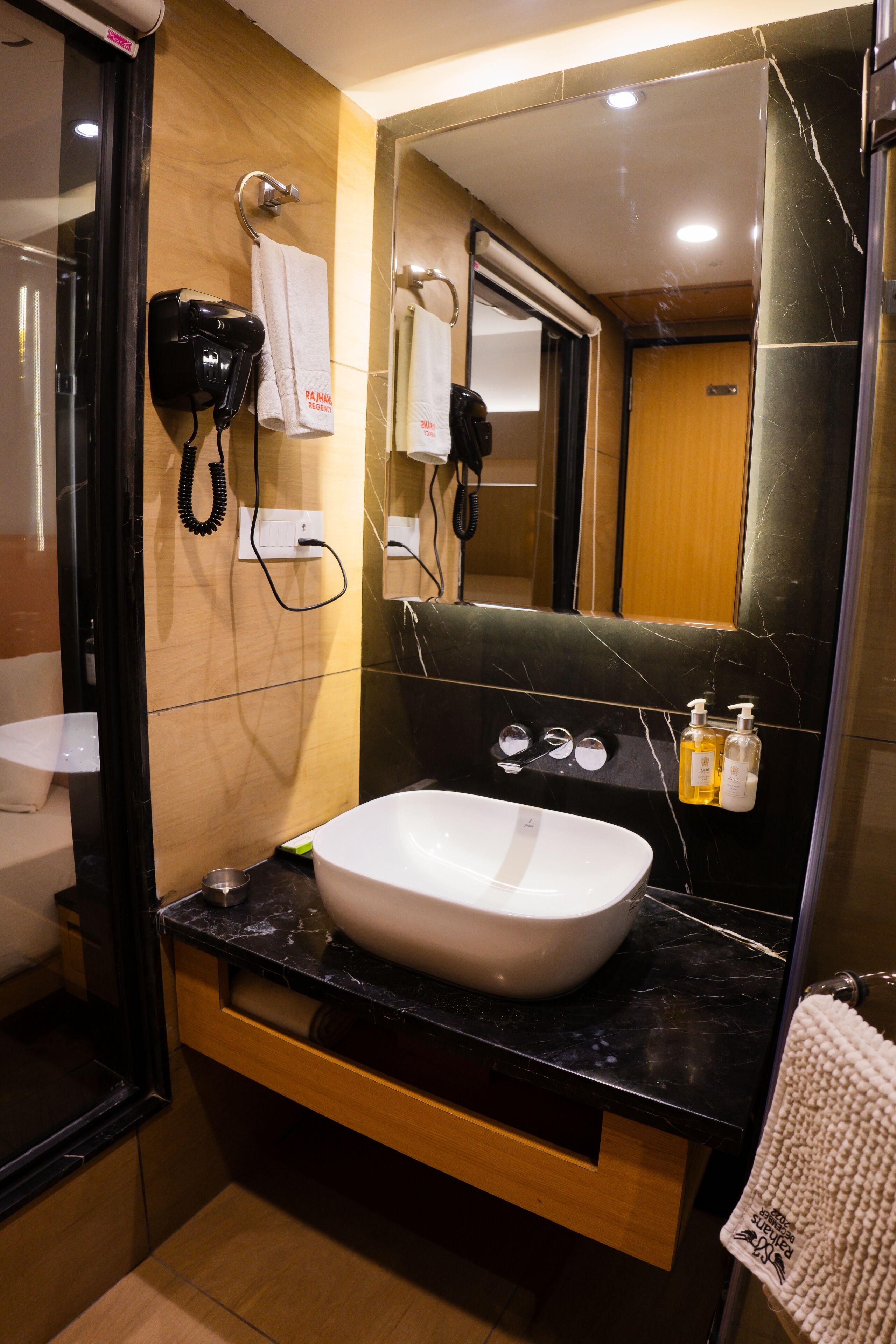 A bathroom with a white sink, black marble countertop, and a mirror above it. There is a hairdryer and phone on the wall, and a towel hanging on the door.