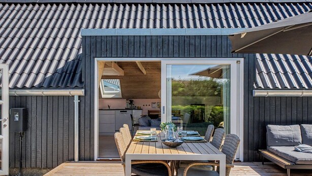 A modern outdoor dining area with a wooden table and chairs, a glass door leading to a kitchen, and a built-in bench.