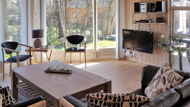 A modern living room with a wooden floor, a sofa, a coffee table, and a TV. There are two chairs, a lamp, and a window with a view of trees and a building.