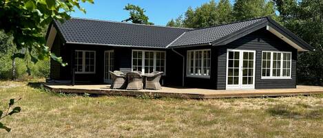Une maison à deux étages, à l’extérieur sombre, dotée de grandes fenêtres et d’un patio couvert aménagé avec du mobilier d’extérieur.