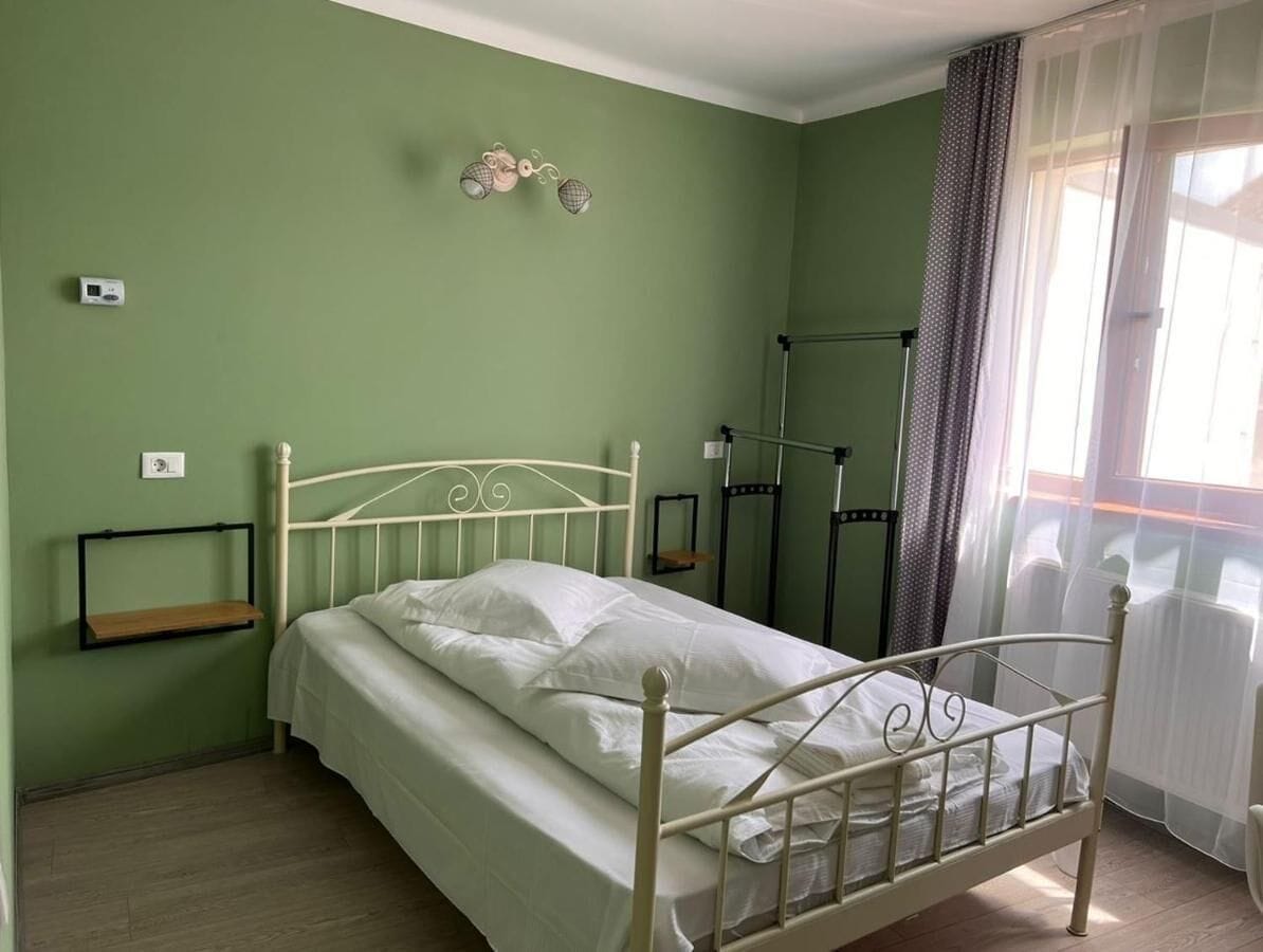 A single-bed room with a metal frame, a bedside table, a shelf, and a window with curtains.