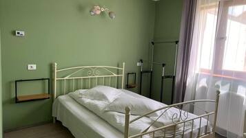 A single-bed room with a metal frame, a bedside table, a shelf, and a window with curtains.