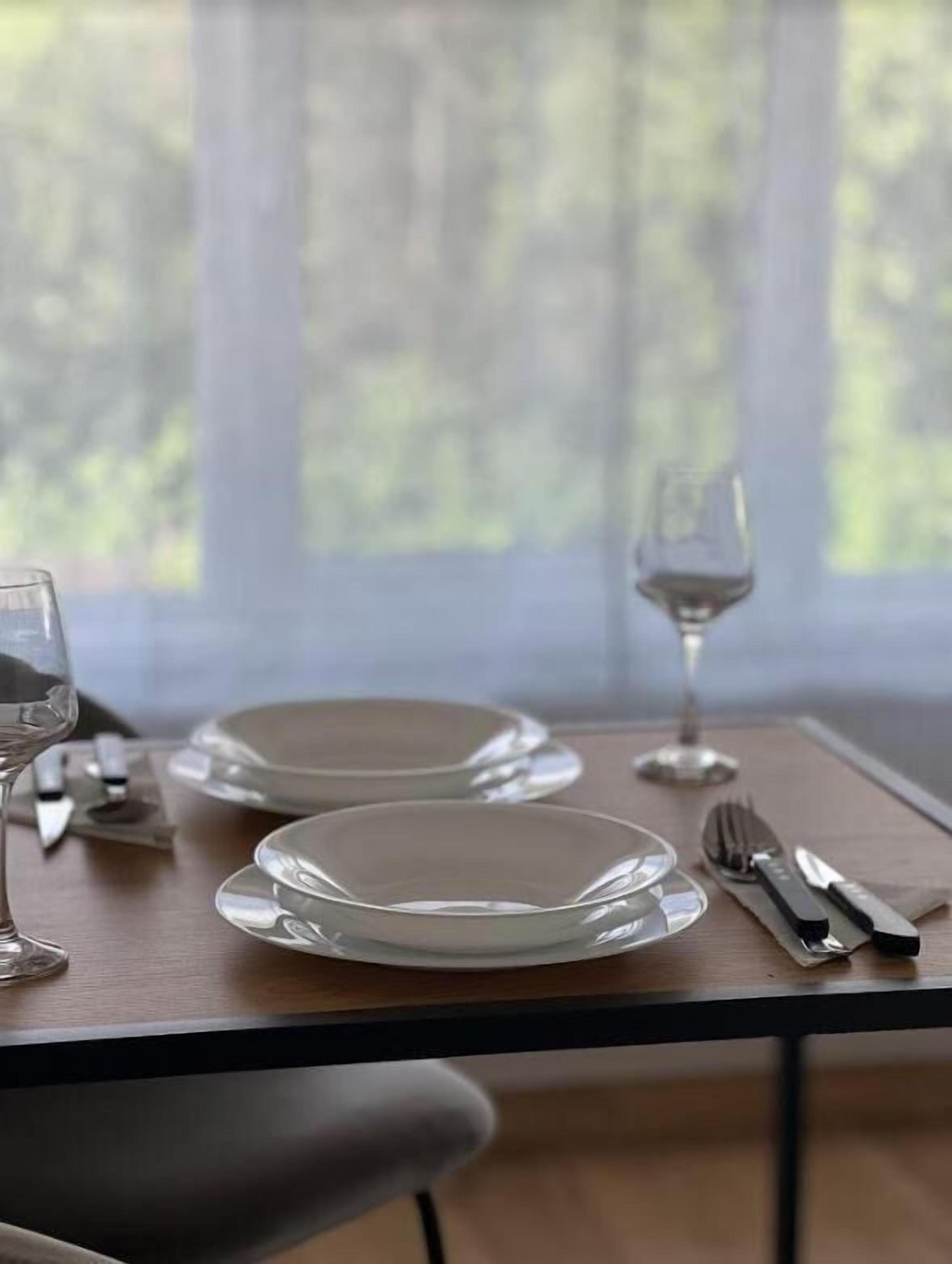 Una mesa de comedor dispuesta con platos, cubiertos y una copa de vino, ubicada cerca de una ventana con vistas a árboles.