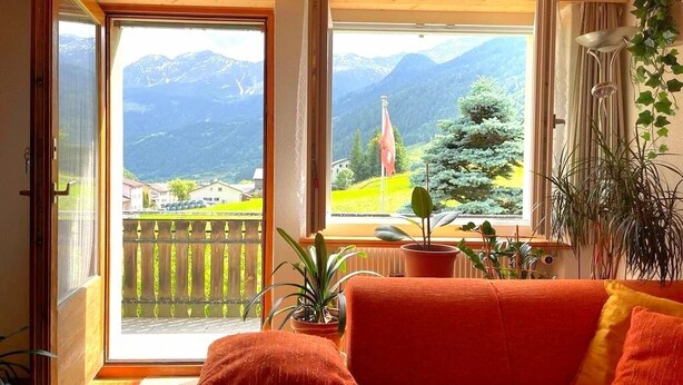 Una habitación con un amplio ventanal que ofrece vistas a la montaña, un sofá rojo y plantas en macetas.