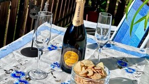 Una mesa con una botella de vino Valdo, dos copas y un bol de aperitivos en un balcón con vistas a un campo verde y un tren.