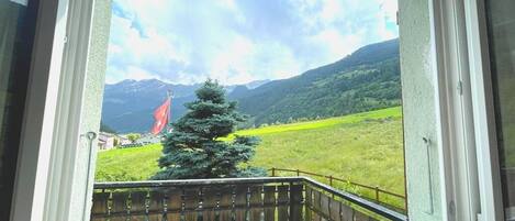 Finestra amb vista a un paisatge verd, un pont de fusta i una serralada de muntanyes al fons.