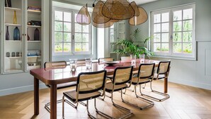 A dining area with a wooden table and six chairs, large windows, and a bookshelf with decorative items.