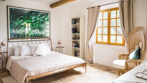 A bedroom with a bed, a bookshelf, a wicker chair, and a large window with curtains.