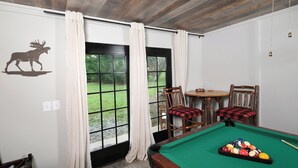Una mesa de billar con superficie de fieltro verde, un vinilo de un alce en la pared y vistas del exterior a través de una puerta con cortinas blancas.