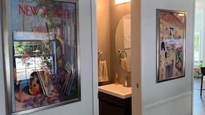 A hallway with framed artwork on the walls, a bathroom with a sink and towel rack, and a door leading to another room.