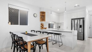 A modern kitchen with white cabinetry, a stainless steel refrigerator, and a dining area with a white table and black chairs. - Spirit of Allura - Located in the beautiful Coolum Beach (Coolum Beach)