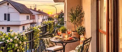 A balcony with a table set for two, surrounded by greenery and a sunset view.