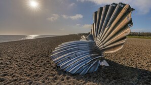 Scultura a forma di guscio metallico su una spiaggia, con il sole sullo sfondo.