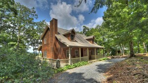 A wooden cabin with a porch, surrounded by trees and a gravel path. - Amber Sunsets dazzles with panoramic views, hot tub luxury, a custom firepit, screened porch, pool table, and pet-friendly hospitality! (Talking Rock)
