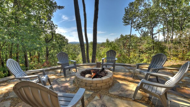 A circular seating area with Adirondack chairs around a fire pit, set on a stone patio with a forest backdrop. - Amber Sunsets dazzles with panoramic views, hot tub luxury, a custom firepit, screened porch, pool table, and pet-friendly hospitality! (Talking Rock)