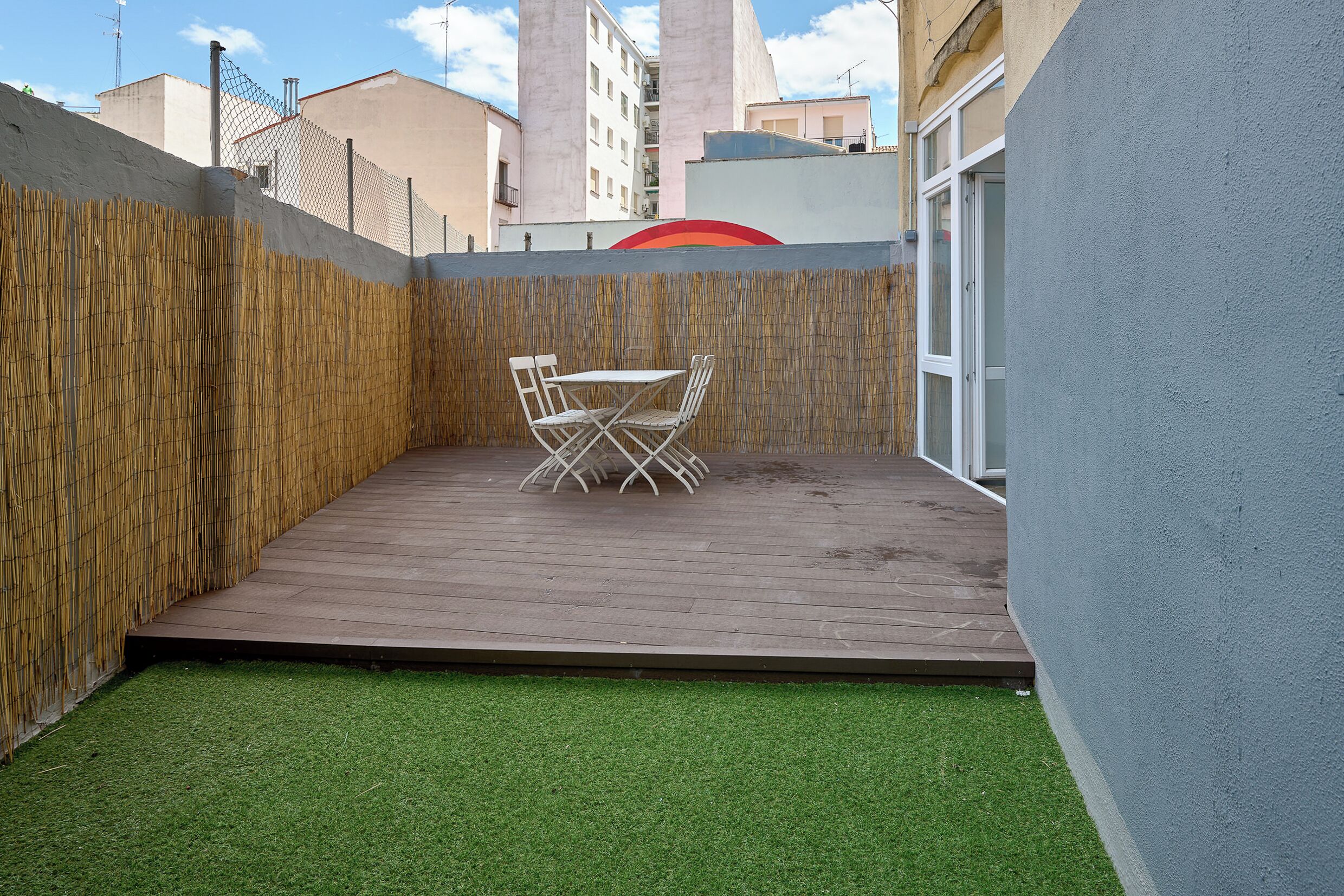 Een klein terras buiten met een tafel en stoelen, omringd door een bamboombordering en een lage muur.