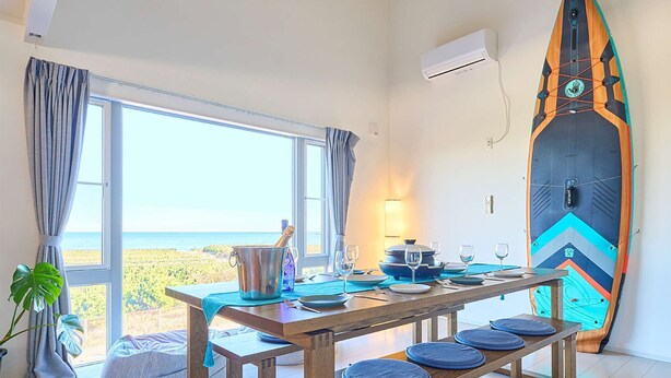 A dining area with a table set for four, a surfboard leaning against the wall, and a view of the ocean through large windows.