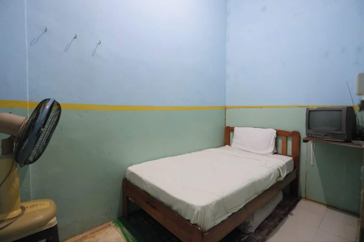 A small, single-bed room with a wooden bed, a fan, a television on a stand, and a yellow stripe on the wall.