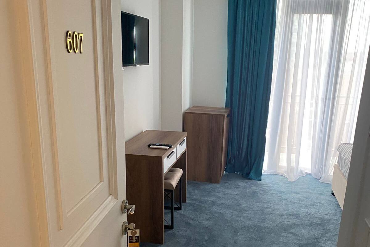 A hotel room with a desk, a television, a bench, and a large window with curtains.