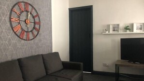 En moderne stue med en grå sofa, et vægur, en dør og et fjernsyn på et stativ.