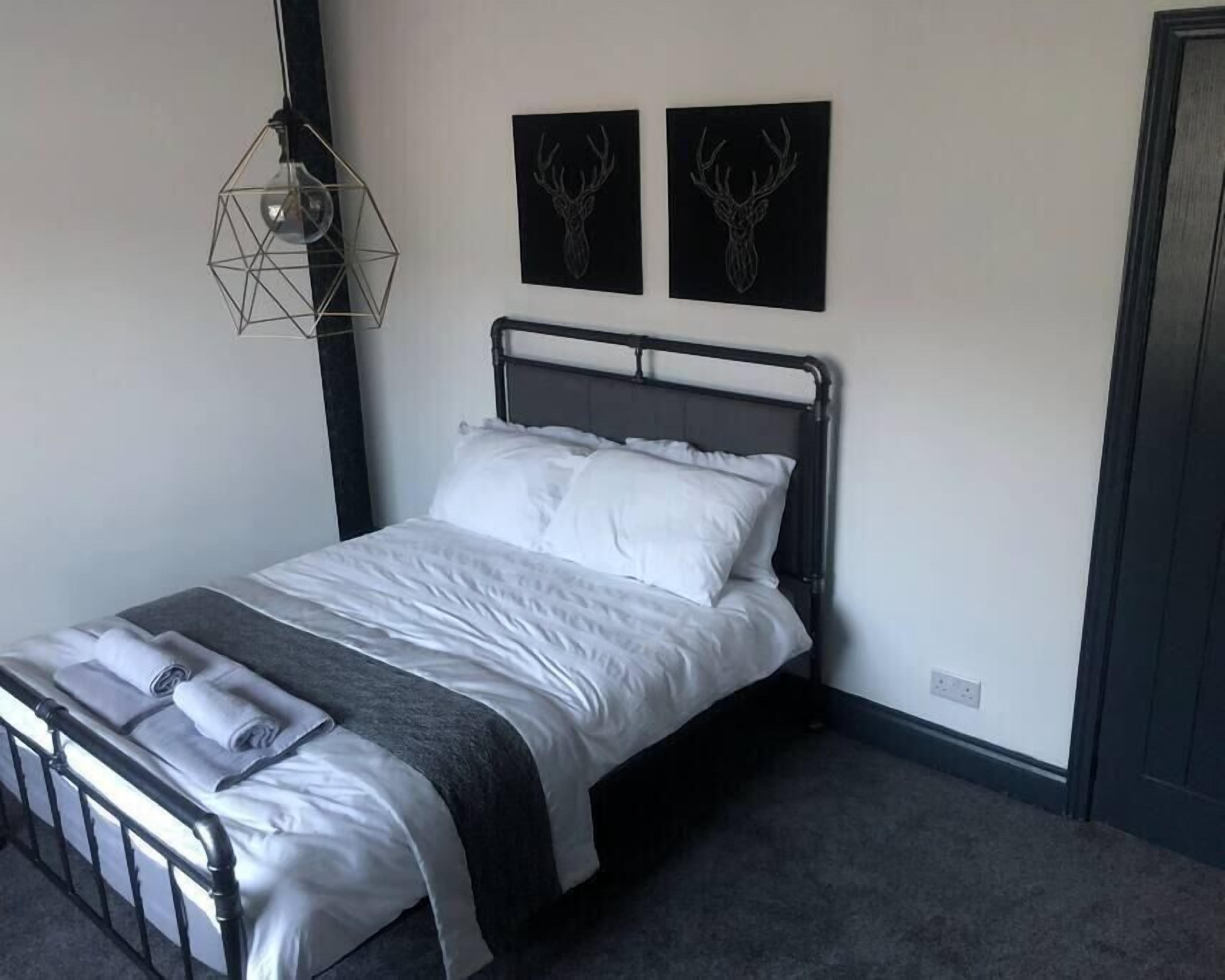 A neatly made bed with white linens and a gray throw blanket, a geometric pendant light, and two wall-mounted deer head art pieces.