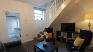 Una sala de estar con sofá, mesa de centro y televisor. Se ve una escalera que sube a otro nivel.