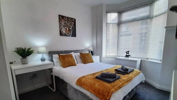 Un dormitorio moderno con una cama grande, una mesita de noche, una planta en maceta, una ventana con persianas y una obra de arte con estampado de leopardo en la pared.
