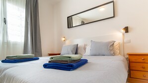 A neatly made bed with folded towels, a bedside table, a nightstand, and a wall-mounted mirror. - Las Casitas de Corralejo (La Oliva)