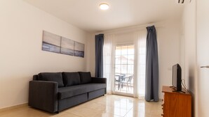 A modern living room with a grey sofa, a wooden TV stand, and a large window with curtains. - Las Casitas de Corralejo (La Oliva)