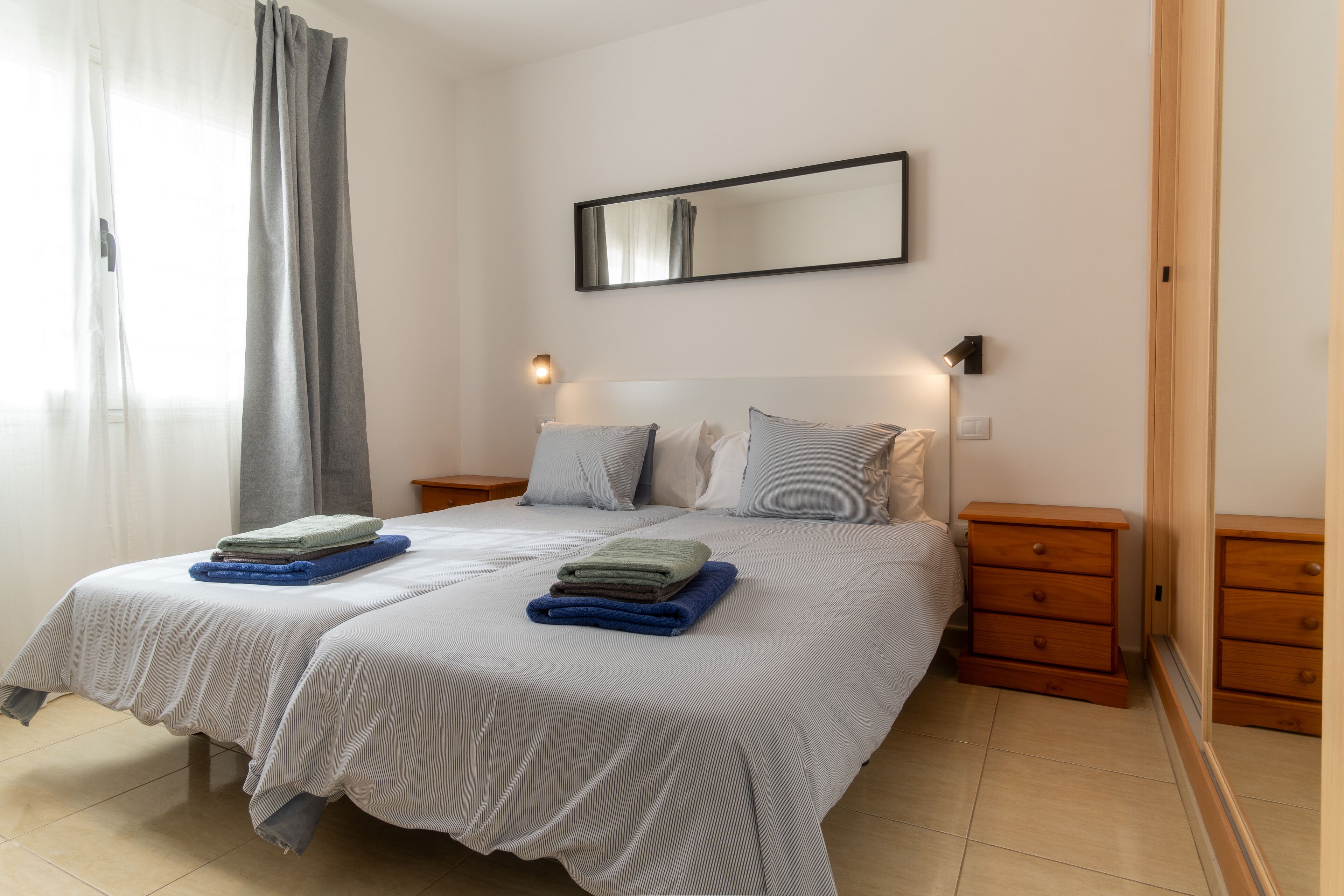 A neatly made bed with two pillows and a folded blanket, a bedside table with a lamp, and a mirror on the wall.