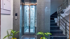 An elevator with glass doors and a decorative panel, a staircase with metal railings, and two potted plants. - DIPLOMAT REGENCY (New Delhi)