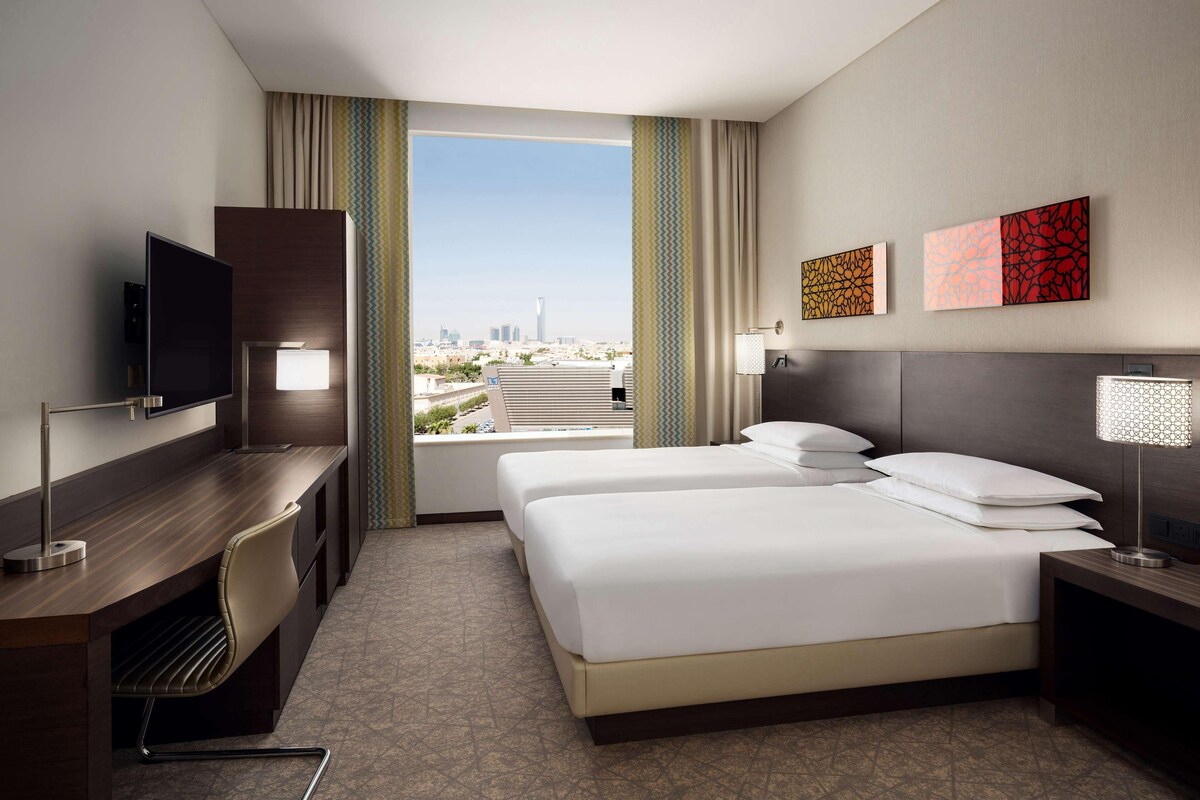 A hotel room with a desk, two beds, a TV, and a city view.
