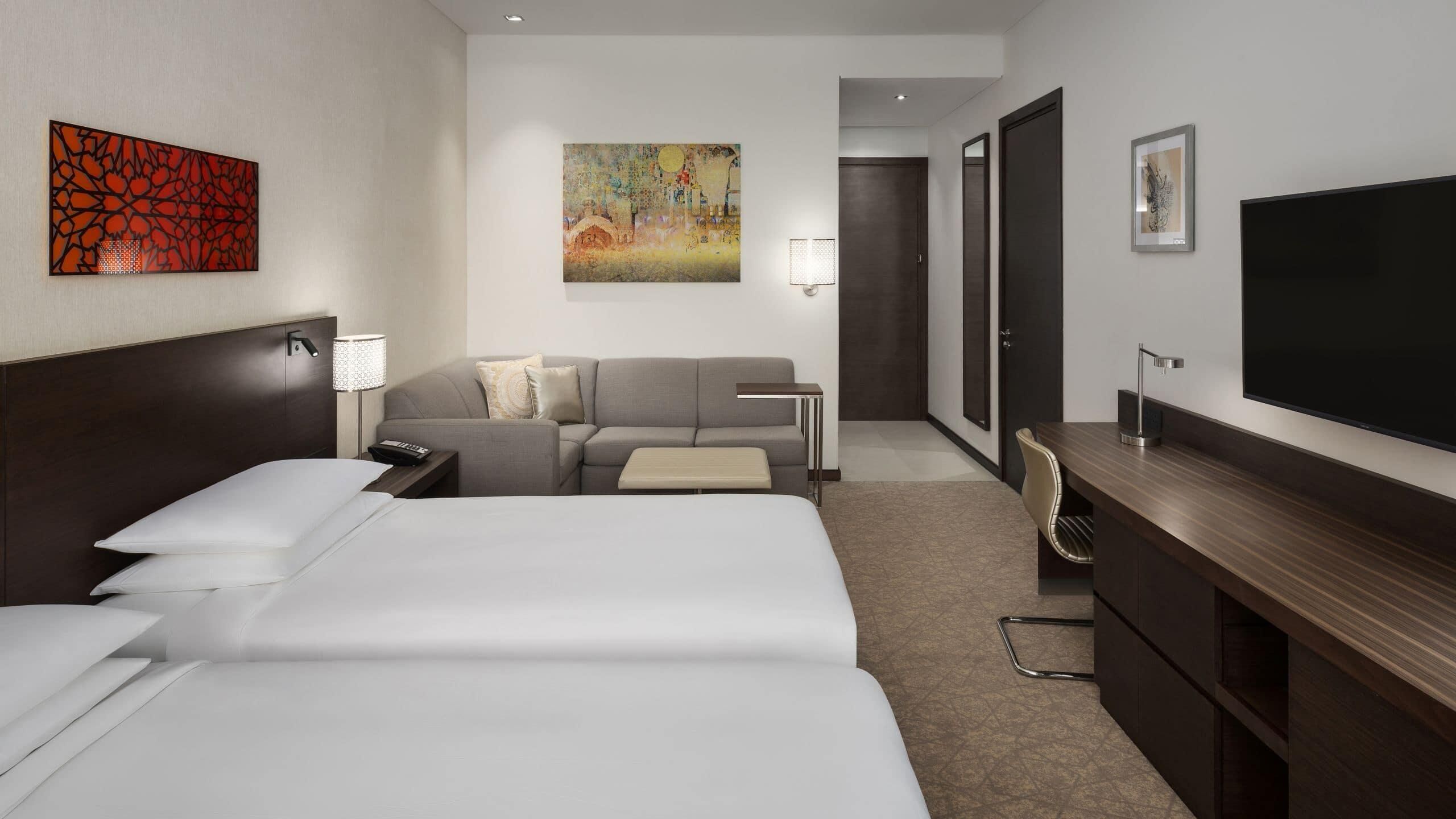 Ein Hotelzimmer mit zwei Betten, einem Schlafsofa, einem Schreibtisch, einem Fernseher und Bildern an den Wänden.